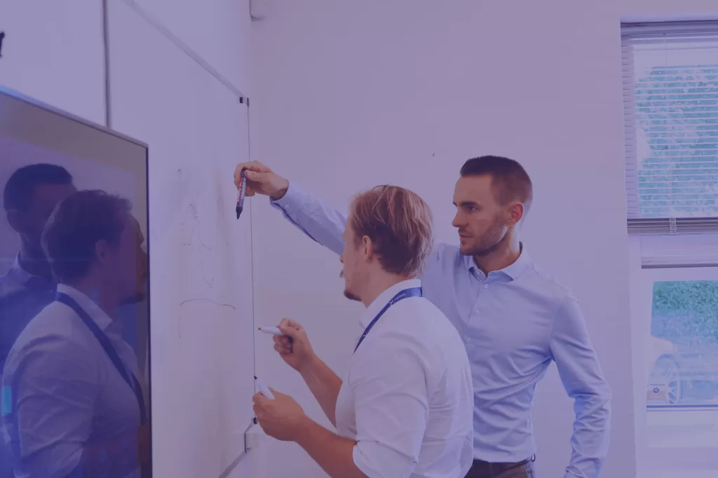Two colleagues collaborating at a whiteboard, discussing diagrams as part of reliable renewable infrastructure planning - purple filter